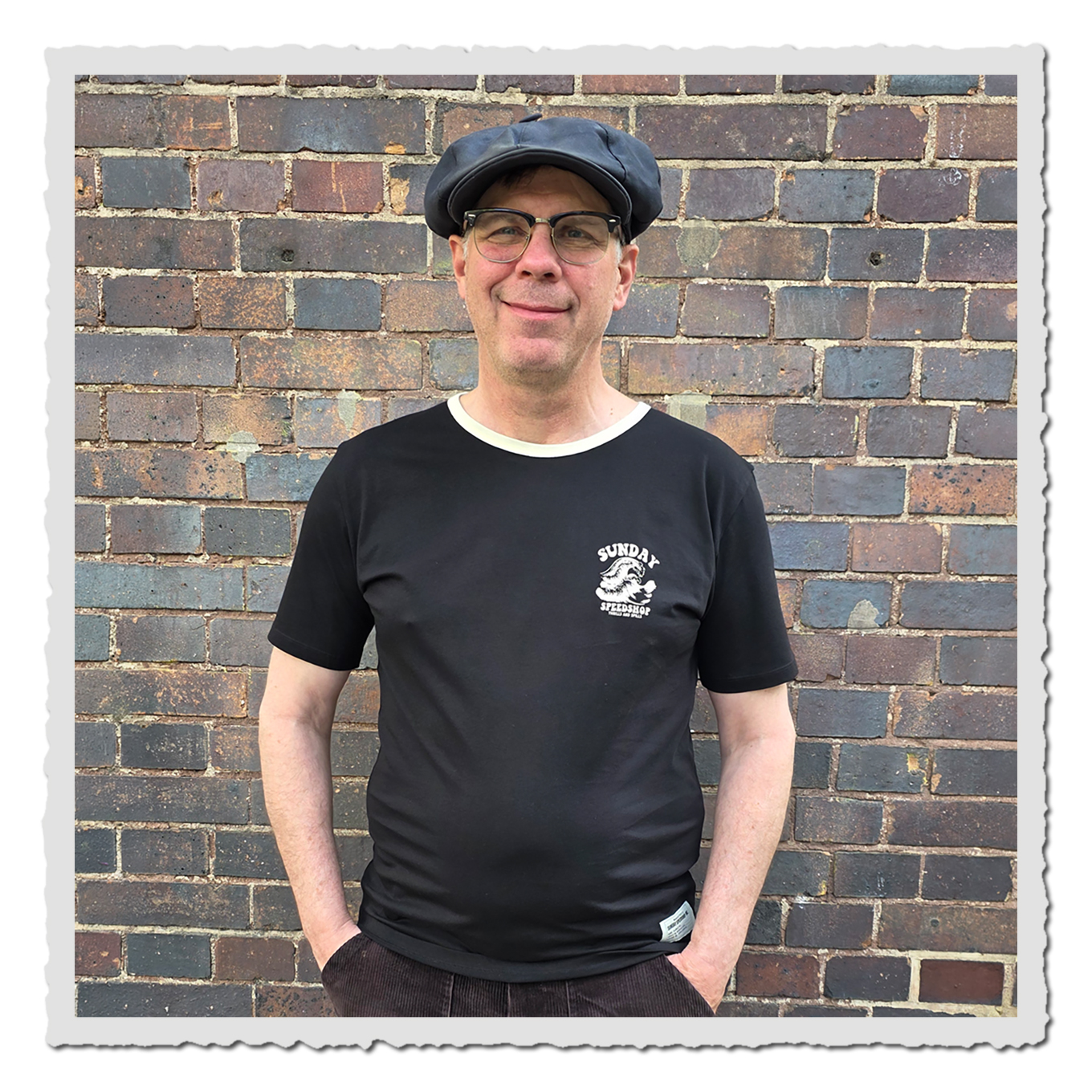 Mann trägt schwarzes T-Shirt mit hellem Brustmotiv vor einer Backsteinwand, Vorderansicht.